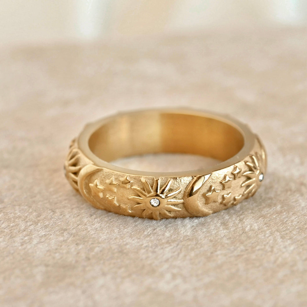 A textured yellow gold band ring with embossed floral and leaf patterns and small clear stones, resting on a soft beige fabric surface.