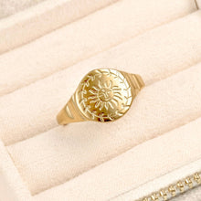 A gold signet ring with an engraved sun and leaf motif, displayed on a cream-colored textured jewelry box.