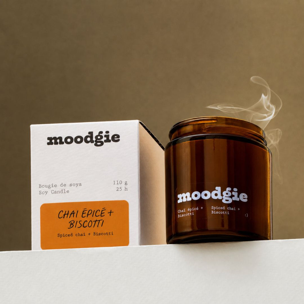A moodégie soy candle in an amber glass jar with visible steam, shown next to its white and orange product box on a clean white surface against a warm taupe background.