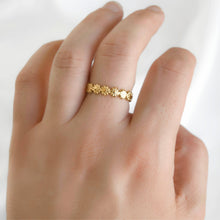 A delicate gold band ring with a row of small flower motifs, worn on the ring finger of a hand against a soft white background.
