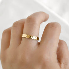A close-up of a hand wearing a thin, hammered gold band ring against a soft, light gray background.