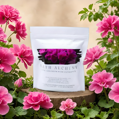A stand-up pouch of Bath Alchemy Rose Geranium Bath Experience sits on a rustic wooden slice surrounded by bright pink roses and green leaves against a soft beige background.