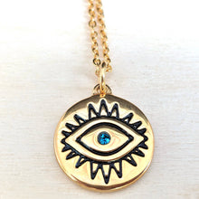 A gold-toned circular pendant necklace featuring an evil eye design with black enamel details and a small blue gemstone at the center, hanging from a gold chain on a light background.