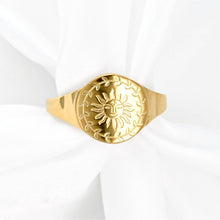 A gold signet ring featuring an embossed sun and leaf wreath design, displayed on a soft white fabric background.