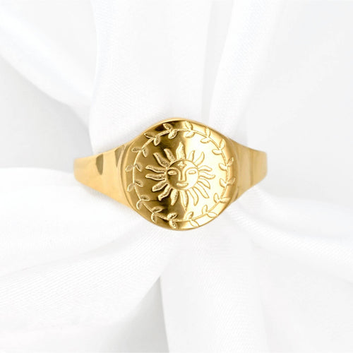 A gold signet ring featuring an embossed sun and leaf wreath design, displayed on a soft white fabric background.