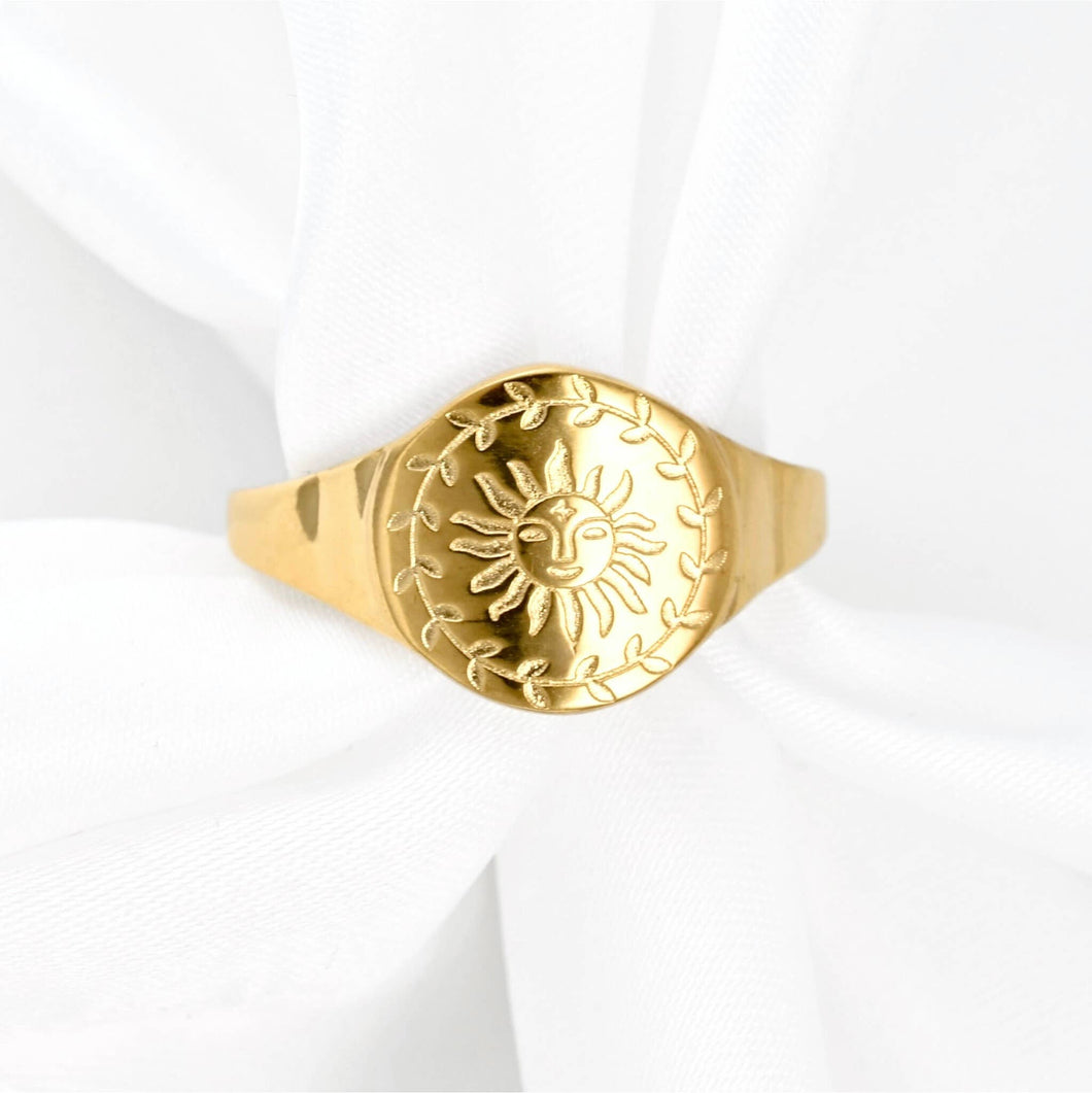 A gold signet ring featuring an embossed sun and leaf wreath design, displayed on a soft white fabric background.