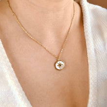 A close-up of a person wearing a delicate gold chain necklace with a small round pendant featuring a starburst design on a white background.