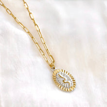 A gold chain necklace with an oval pendant featuring a sunburst border and a central white inlay with a gold bird motif, displayed on a soft white fabric background.