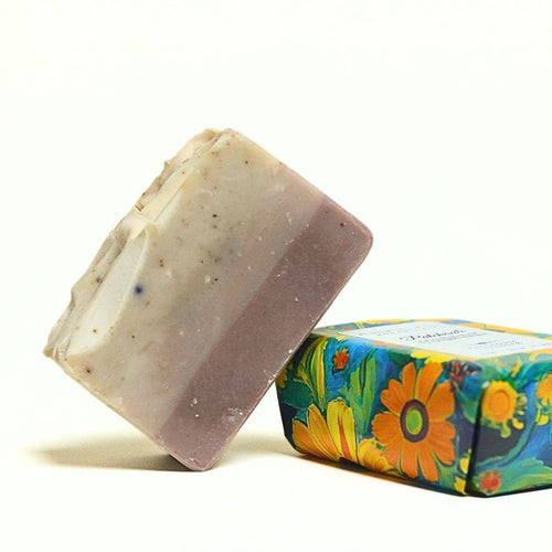 A handmade rectangular bar of soap with a lavender-to-cream gradient and small dark specks, standing next to its colorful floral-patterned paperboard packaging box on a white background.