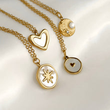 Three gold-tone pendant necklaces with white inlay details, displayed on a soft white satin background. The necklaces feature heart, sunburst, and heart-with-heart pendants.