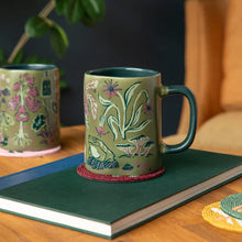 A ceramic mug with an olive green base and hand-painted nature illustrations of a frog, bird, and plants, styled on a dark green book with a matching second mug in the background.