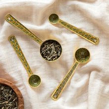 Gold and green patterned spoons with a small bowl containing tea leaves on a beige fabric background.