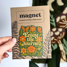 Cardboard magnet with floral design and text 'Enjoy the Journey' held by a hand.