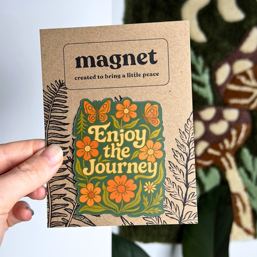 Cardboard magnet with floral design and text 'Enjoy the Journey' held by a hand.