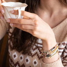 Person holding a small white ceramic cup with a patterned sweater and bracelets.