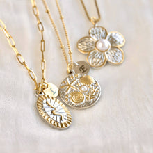 Three gold and silver necklaces with textured charms are arranged on a soft white fabric background. One necklace has a flower-shaped charm with a pearl center, another has a round sunburst charm, and the third has a round shell-and-sea-themed charm with a small pearl.