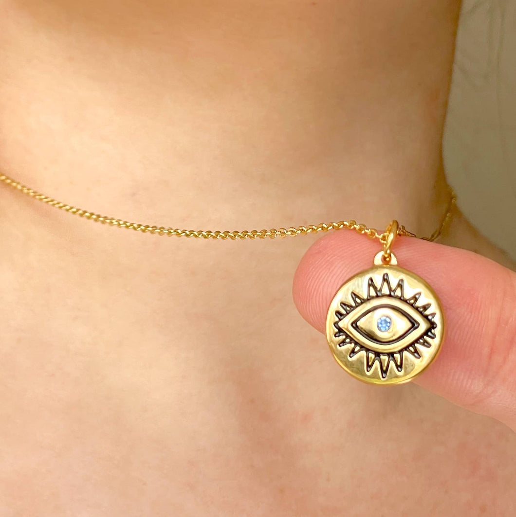 A gold-toned chain necklace with a circular evil eye pendant worn on a person's neck, with a finger holding the pendant in front of the face.