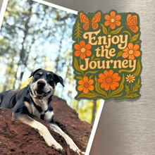 Dog lying on the ground with a decorative magnet reading 'Enjoy the Journey' featuring flowers and butterflies.