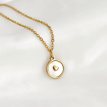 A delicate gold chain necklace with a round pendant featuring a white inlay and a small gold heart in the center, displayed on a soft white fabric background.