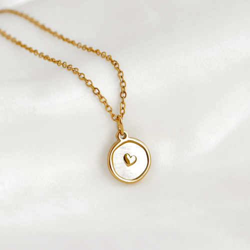 A delicate gold chain necklace with a round pendant featuring a white inlay and a small gold heart in the center, displayed on a soft white fabric background.