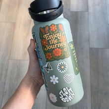 Hand holding a green water bottle with floral design and text on a wooden floor background