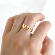 A close-up of a hand wearing a delicate gold ring with intricate cutout floral patterns, set against a soft, bright white background.