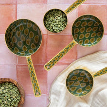 Three green and gold patterned ceramic measuring cups with handles are arranged on a pink tiled surface, with green beans in bowls and a cream cloth nearby.