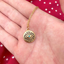A gold-toned evil eye pendant necklace with a small turquoise-blue stone in the center, resting on an open hand over a red and white polka dot fabric background.
