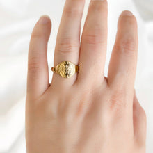 A close-up of a hand wearing a textured gold signet ring with a lion head motif, set against a soft white fabric background.