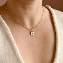 A close-up of a person wearing a delicate gold chain necklace with a small round pendant featuring a white inlay and a tiny gold heart.