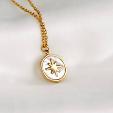 A gold chain necklace with a round wax-seal style pendant featuring a white mother-of-pearl background and a gold starburst symbol, displayed on a soft white fabric surface.