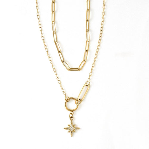 A gold layered necklace featuring a paperclip chain, a link chain, a ring connector, and a small starburst pendant with a clear central stone, shown on a white background.