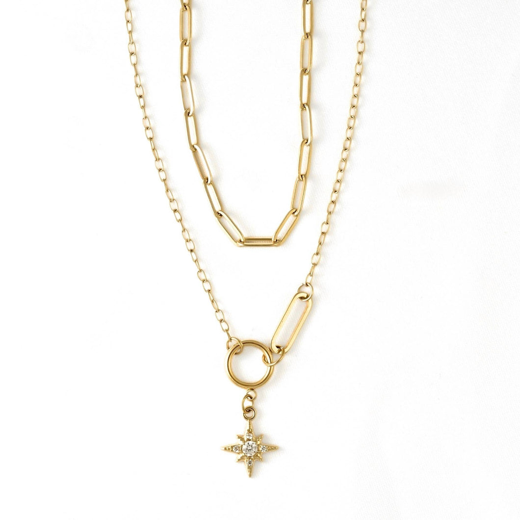 A gold layered necklace featuring a paperclip chain, a link chain, a ring connector, and a small starburst pendant with a clear central stone, shown on a white background.