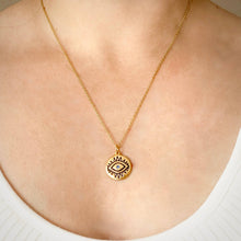 A gold chain necklace with a circular pendant featuring an eye design, worn on a person's neck over a white ribbed top.