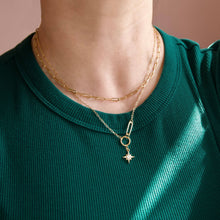 A close-up of a person wearing a double-layered gold necklace. The top layer features a paperclip-shaped pendant with a small star charm dangling below it, worn over a dark green ribbed t-shirt.