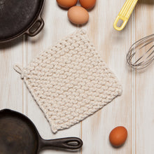 Textured pot holder on a wooden surface with eggs and a whisk.
