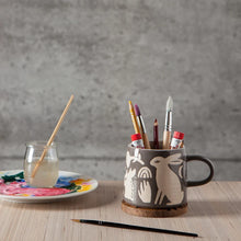 A dark gray ceramic mug with a beige rabbit pattern sits on a cork coaster on a wooden table, filled with paintbrushes and art supplies. To the left is a glass of water with a paintbrush and a painted plate, set against a textured gray wall.